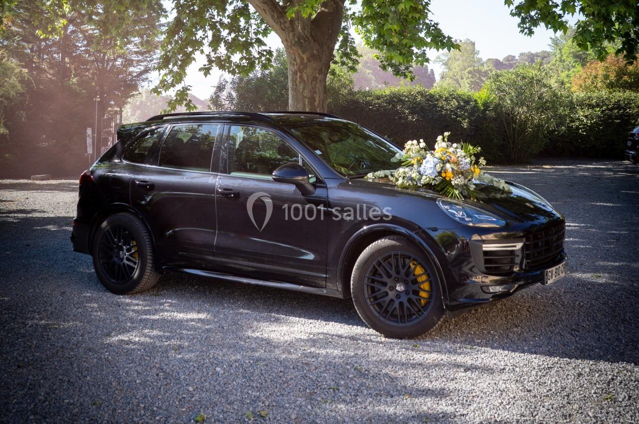Voiture noire décorée de fleurs blanches et jaunes, stationnée sur un chemin gravillonné sous des arbres.