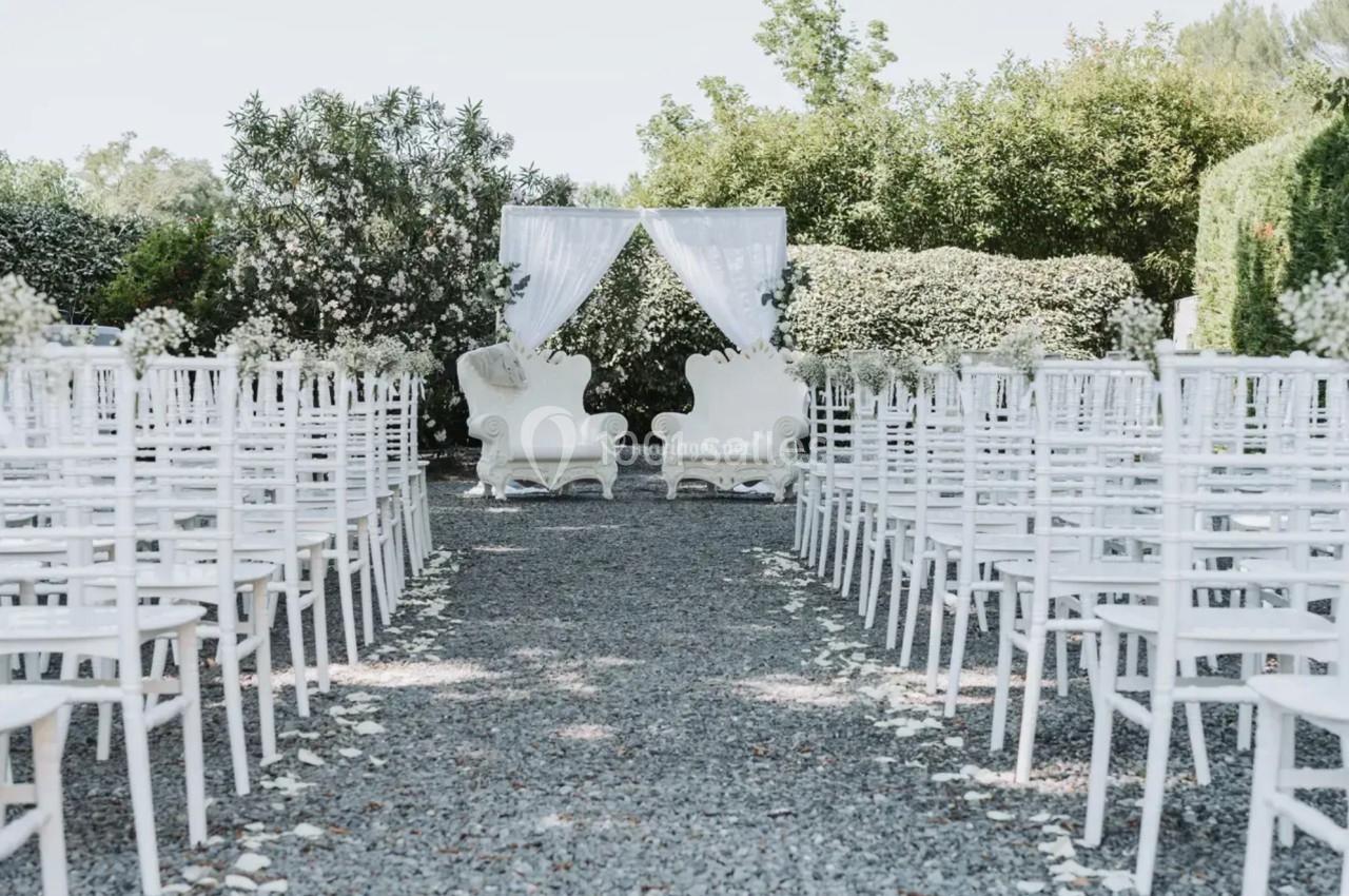 Allée centrale bordée de chaises blanches menant à une arche décorée de rideaux blancs dans un jardin.