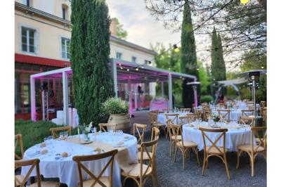 Grande table dressée en extérieur avec des chaises en bois, sous une pergola illuminée de lumières violettes, devant un…