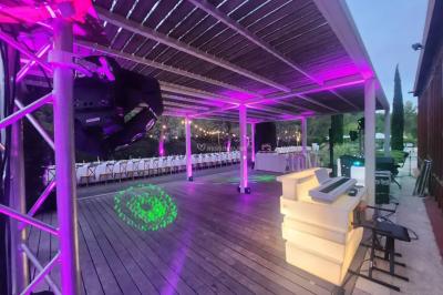 Grande table dressée en extérieur avec des chaises en bois, sous une pergola illuminée de lumières violettes, devant un…