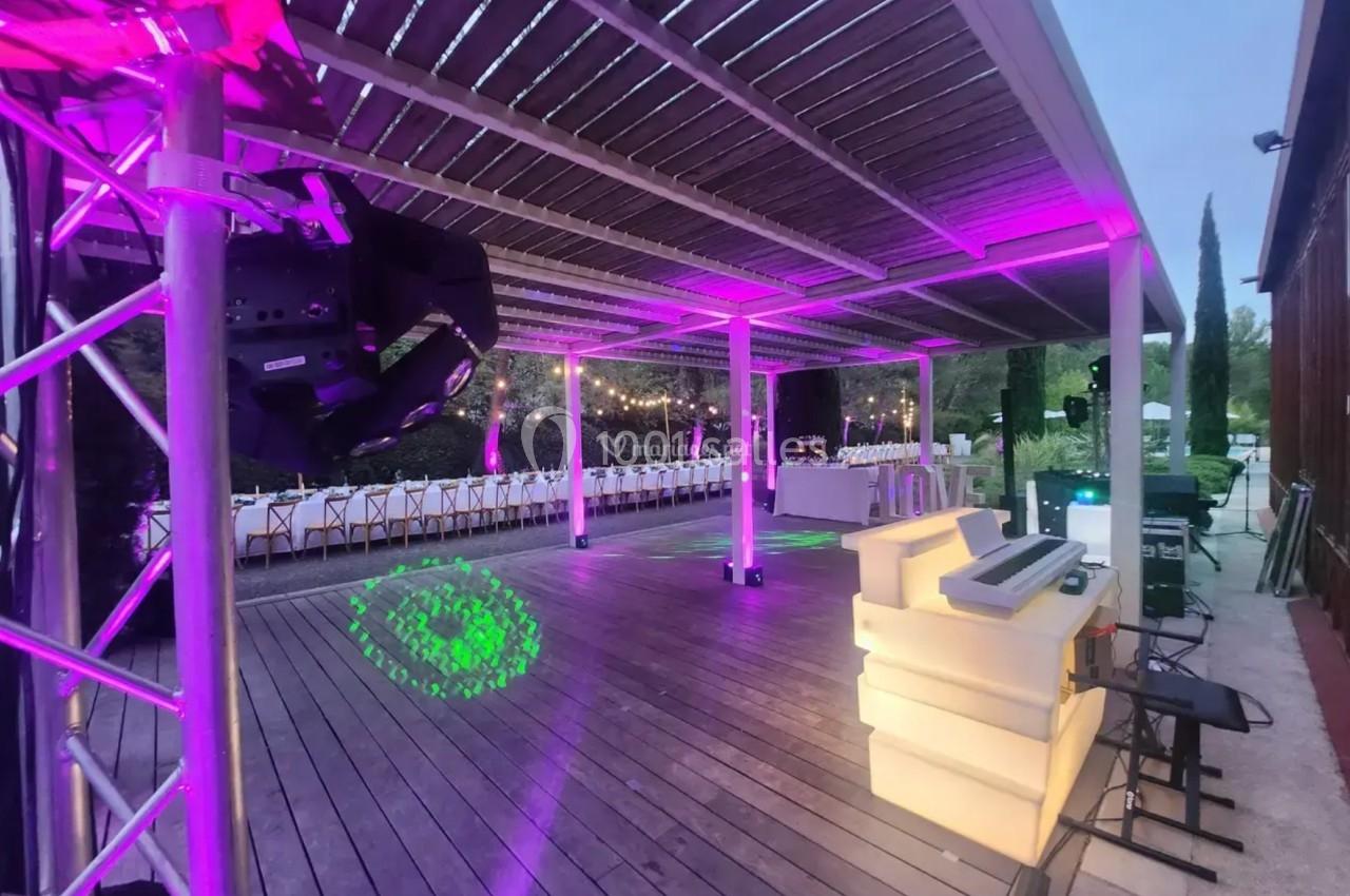 Terrasse en bois éclairée par des lumières colorées, avec tables dressées et espace DJ en arrière-plan.