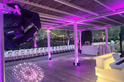 Grande table dressée en extérieur avec des chaises en bois, sous une pergola illuminée de lumières violettes, devant un…