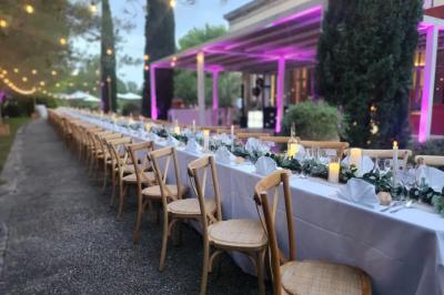 Grande table dressée en extérieur avec des chaises en bois, sous une pergola illuminée de lumières violettes, devant un…