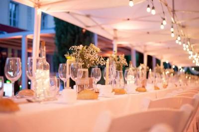 Grande table dressée en extérieur avec des chaises en bois, sous une pergola illuminée de lumières violettes, devant un…