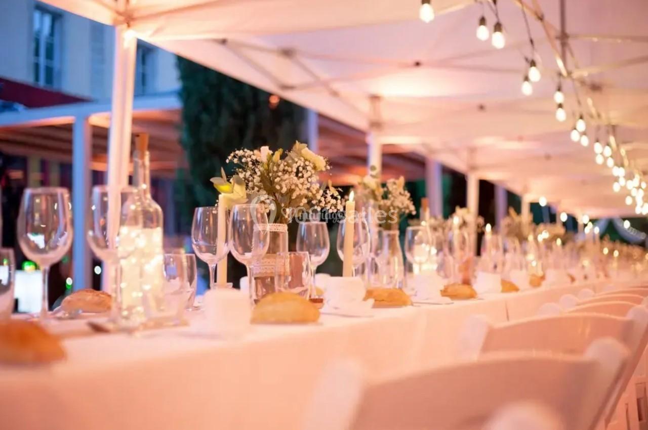 Table élégamment dressée en extérieur sous des guirlandes lumineuses, avec fleurs, verres et pains disposés.