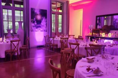 Grande table dressée en extérieur avec des chaises en bois, sous une pergola illuminée de lumières violettes, devant un…