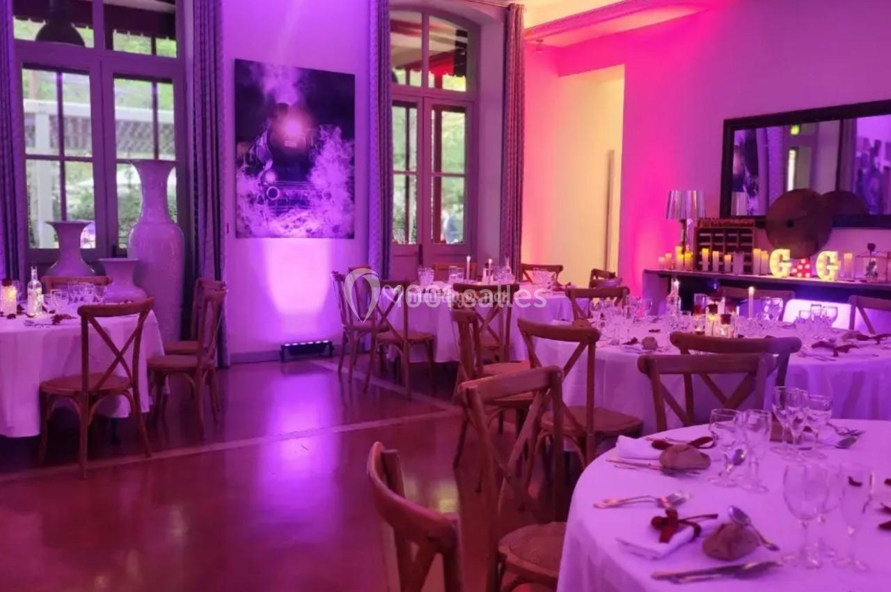 Salle de réception élégante avec tables rondes dressées, éclairage tamisé rose et violet, grandes fenêtres en arrière-plan.