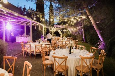Grande table dressée en extérieur avec des chaises en bois, sous une pergola illuminée de lumières violettes, devant un…