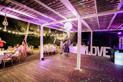 Grande table dressée en extérieur avec des chaises en bois, sous une pergola illuminée de lumières violettes, devant un…