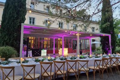 Grande table dressée en extérieur avec des chaises en bois, sous une pergola illuminée de lumières violettes, devant un…
