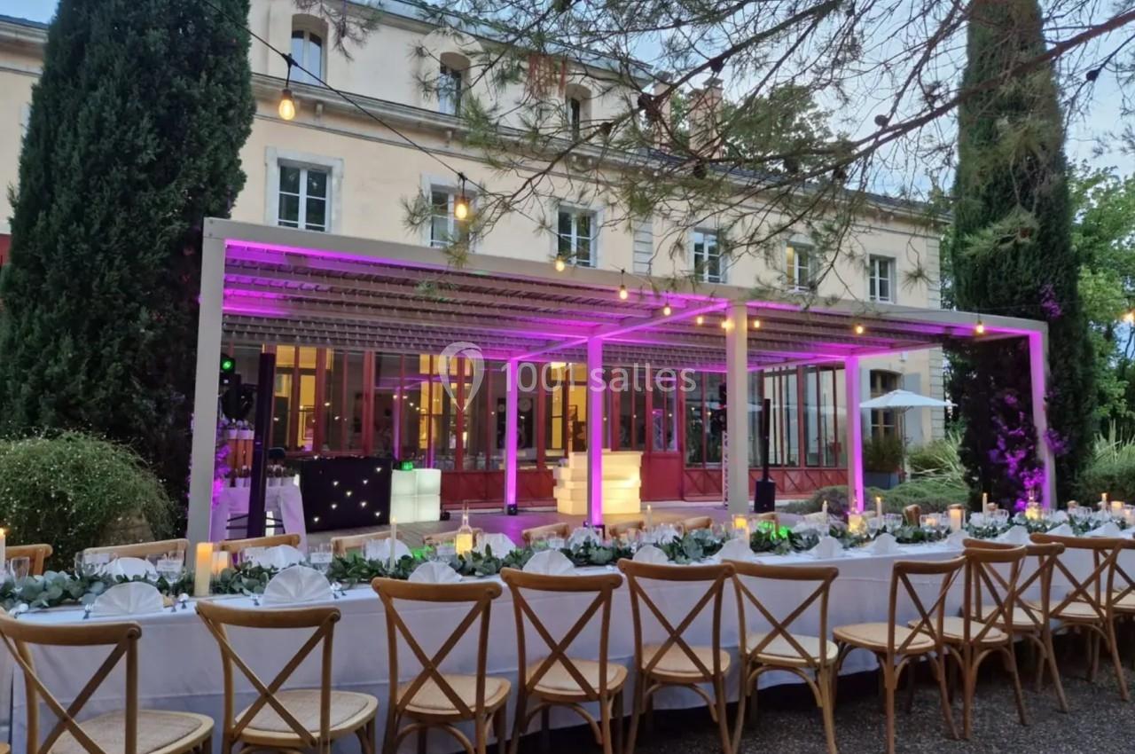 Grande table dressée en extérieur avec des chaises en bois, sous une pergola illuminée de lumières violettes, devant un…