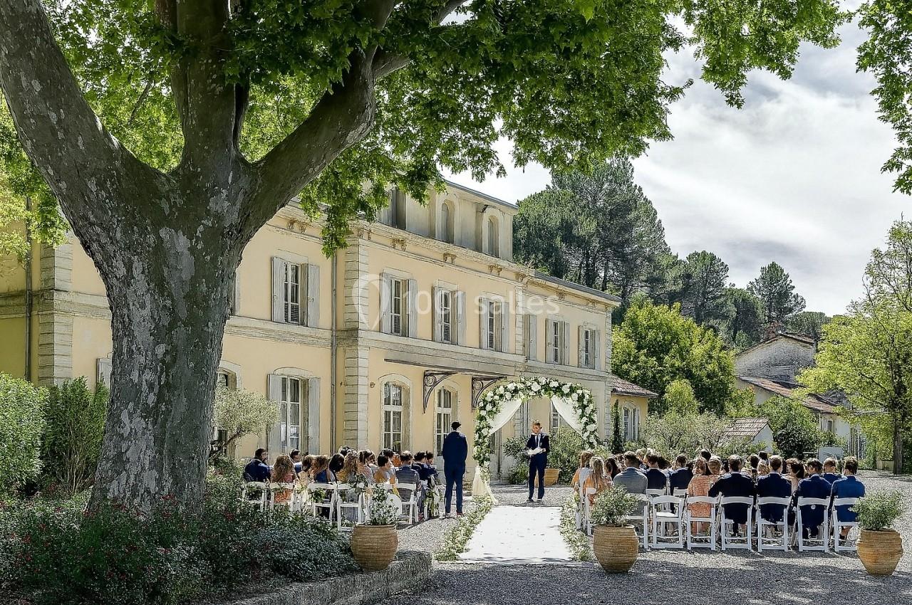 Cérémonie de mariage en plein air devant une grande bâtisse jaune, avec des invités assis sous un arbre.
