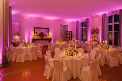 Grande table dressée en extérieur avec des chaises en bois, sous une pergola illuminée de lumières violettes, devant un…