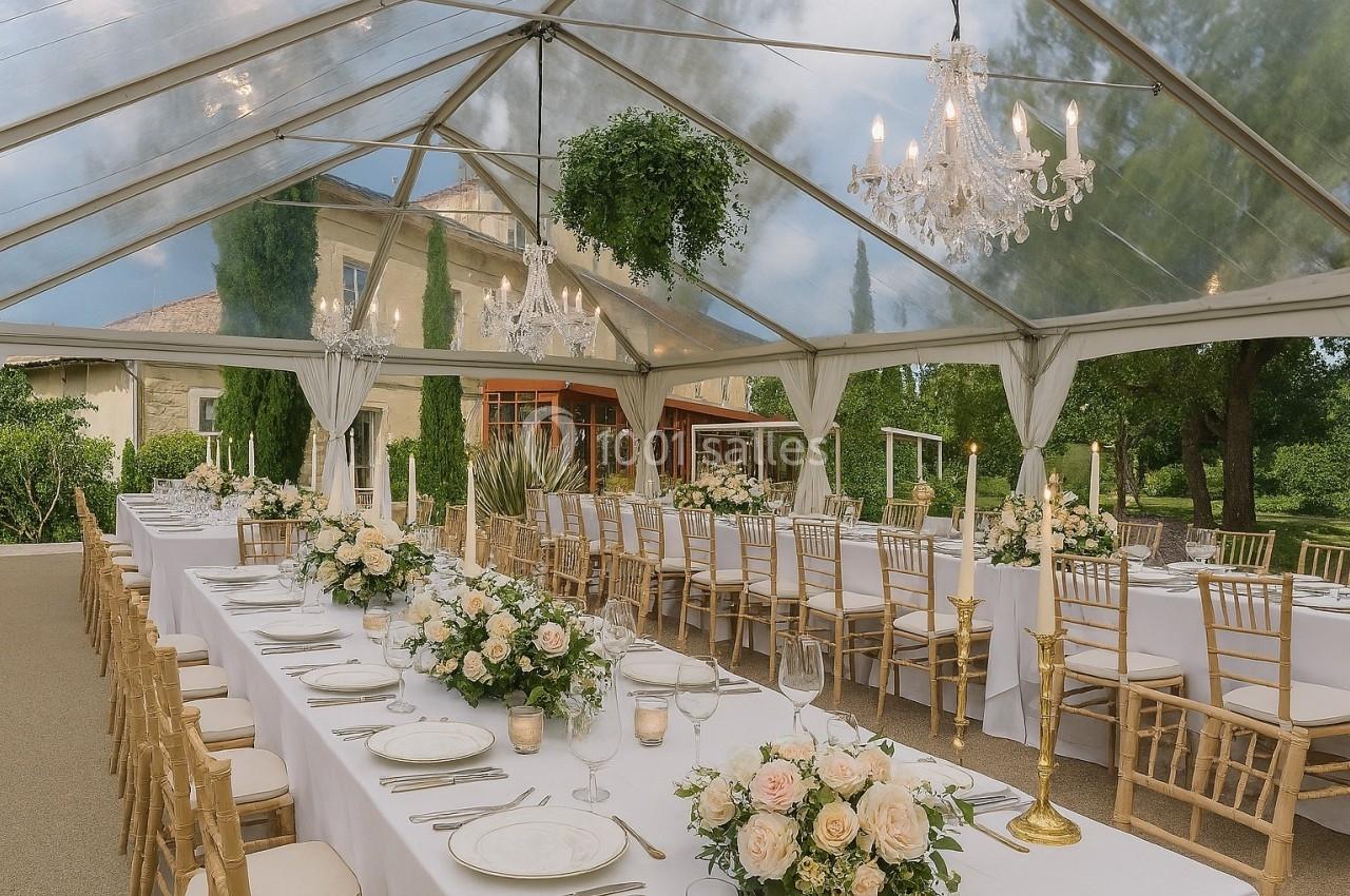 Tables élégamment dressées sous une tente transparente avec chandeliers, fleurs blanches et chaises dorées.