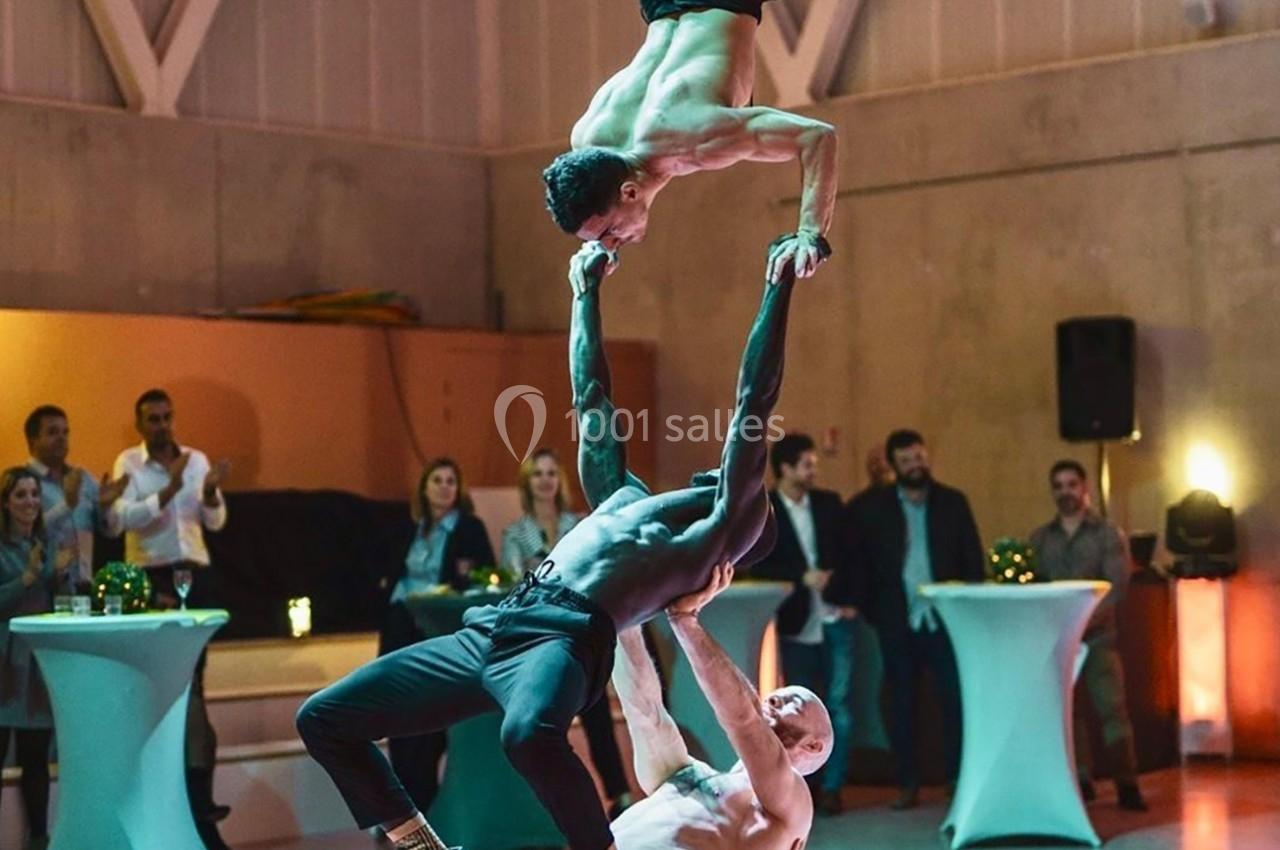 Trois acrobates réalisent une figure d'équilibre dans une salle avec des spectateurs en arrière-plan.