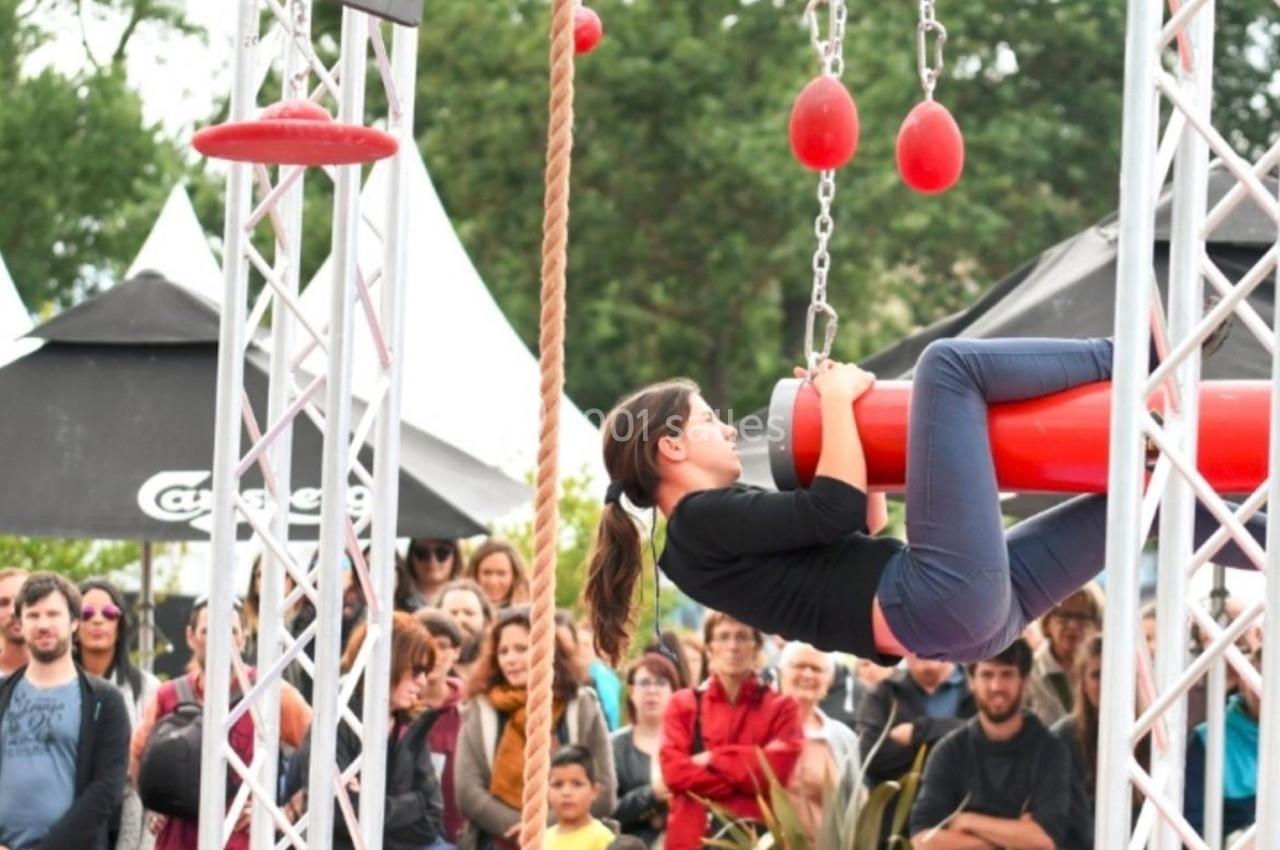 Une femme grimpe sur une structure d'obstacles rouges lors d'un événement en plein air, devant un public attentif.