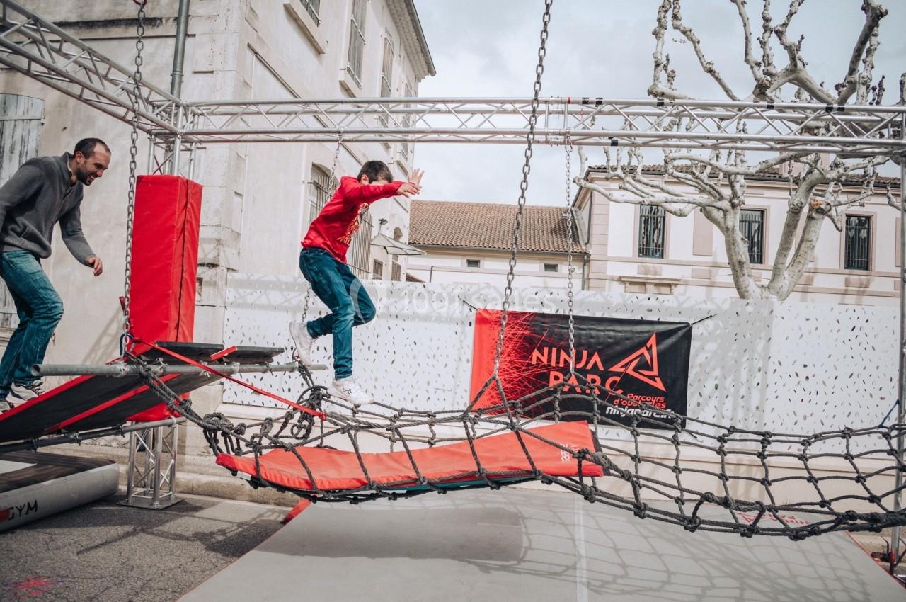 Un enfant traverse un pont de cordes dans un parcours d'obstacles en plein air, sous la surveillance d'un adulte.