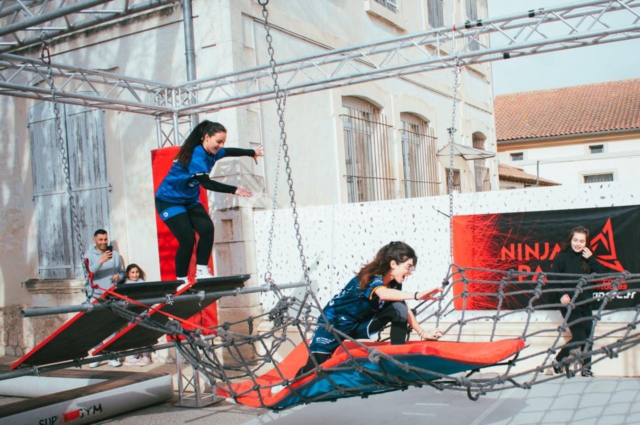 Deux femmes franchissent un parcours d'obstacles avec des filets et des plateformes en extérieur.