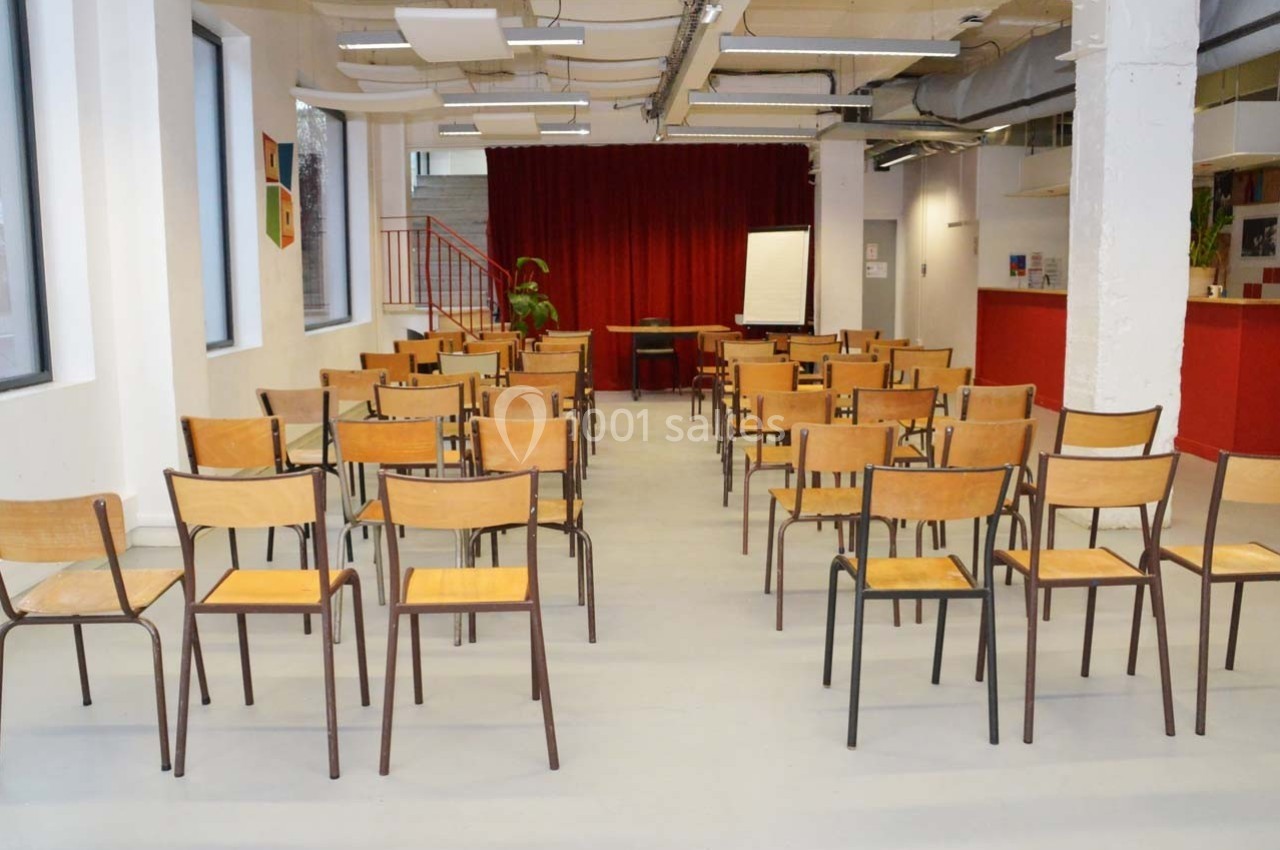 Salle lumineuse avec des rangées de chaises en bois disposées face à un tableau blanc et un rideau rouge.