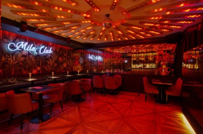 Salle de restaurant élégante avec éclairage tamisé, tables rondes, chaises rembourrées et bar en arrière-plan.