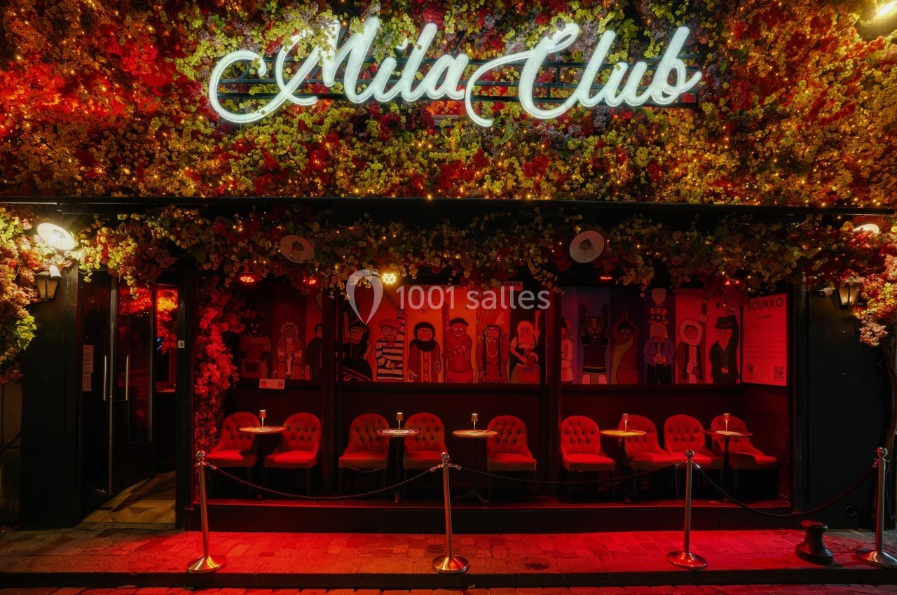 Façade d'un club décorée de fleurs lumineuses, avec enseigne néon et terrasse équipée de chaises et tables.