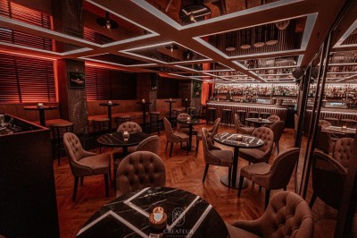 Salle de restaurant élégante avec éclairage tamisé, tables rondes, chaises rembourrées et bar en arrière-plan.