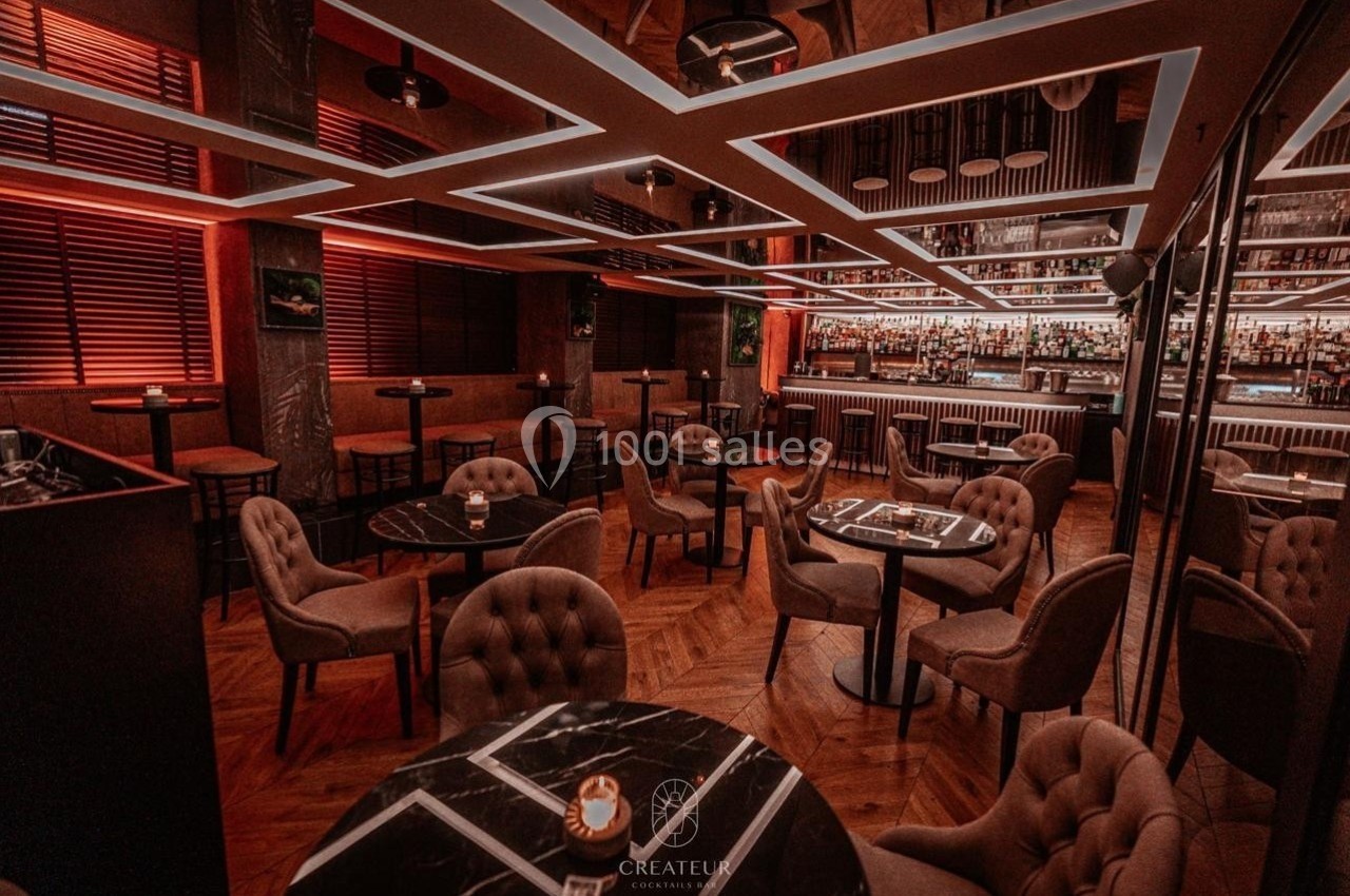 Salle de restaurant élégante avec éclairage tamisé, tables rondes, chaises rembourrées et bar en arrière-plan.