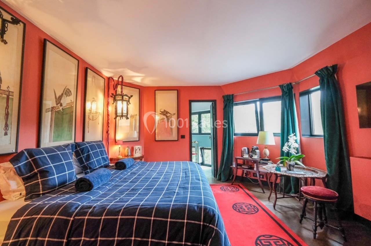 Chambre lumineuse avec murs rouges, lit à motif à carreaux, décorations asiatiques et grandes fenêtres avec rideaux verts.