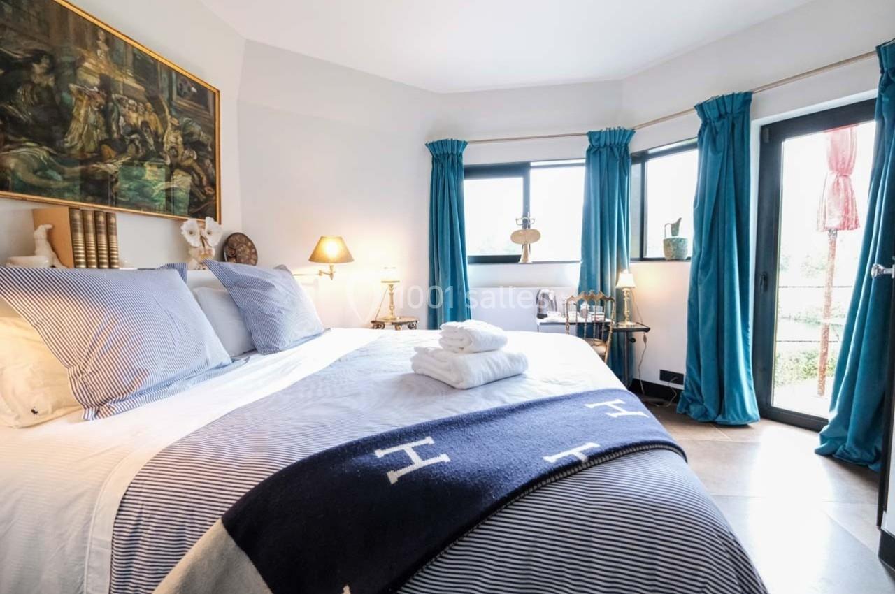 Chambre lumineuse avec lit double, draps rayés, rideaux bleus, tableau mural et accès à un balcon.