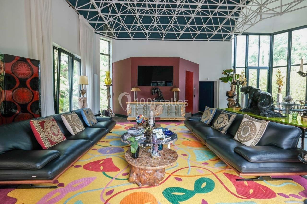 Salon spacieux avec canapés noirs, tapis coloré, meubles éclectiques et grandes fenêtres laissant entrer la lumière…