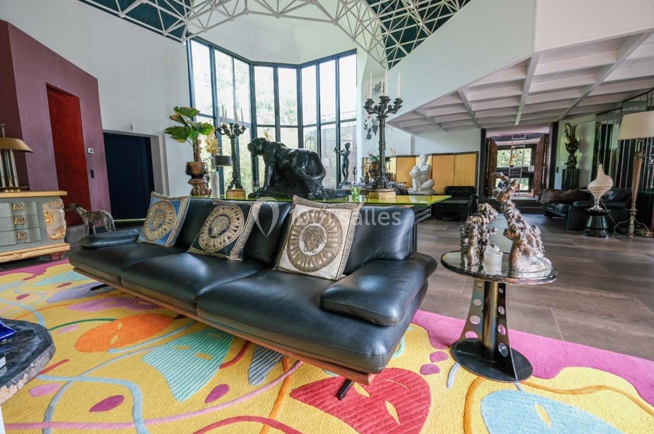 Salon moderne avec canapé en cuir noir, coussins décoratifs, tapis coloré et grandes baies vitrées.