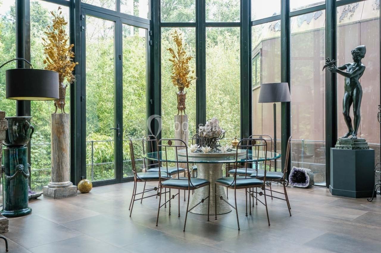 Salle à manger lumineuse avec table ronde en verre, chaises métalliques et grandes baies vitrées donnant sur un jardin…