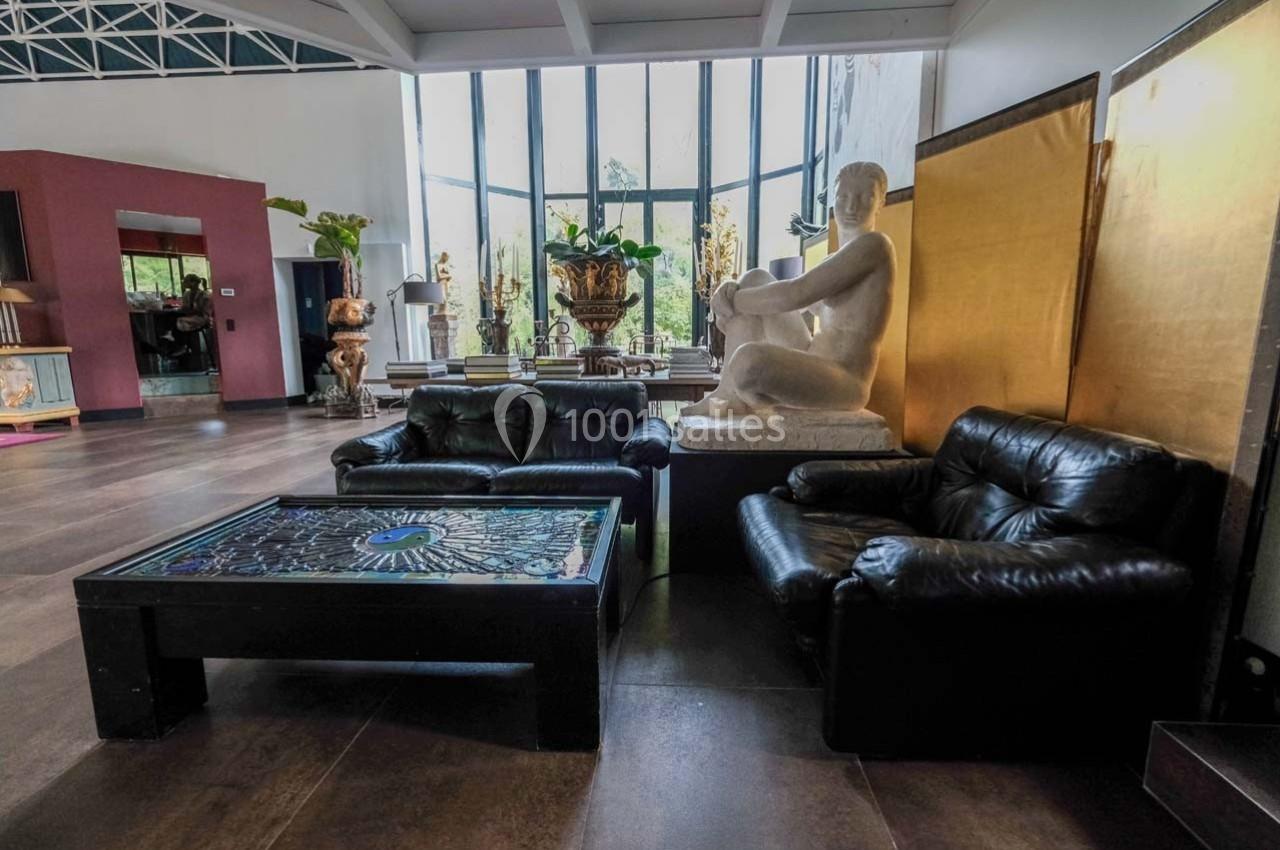 Salon avec deux fauteuils en cuir noir, une table basse en verre décoré et une statue blanche près d'une grande baie vitrée.