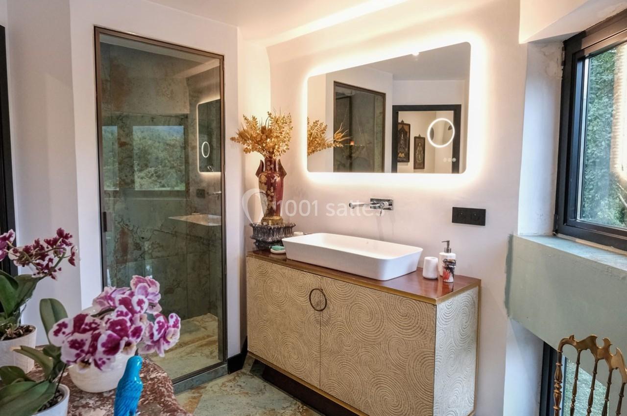 Salle de bain moderne avec vasque blanche, miroir éclairé, douche vitrée et décoration florale.