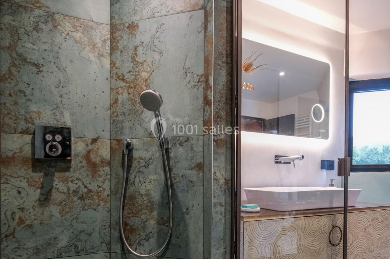 Douche moderne avec carrelage texturé et vue partielle sur un lavabo et un miroir dans une salle de bain lumineuse.