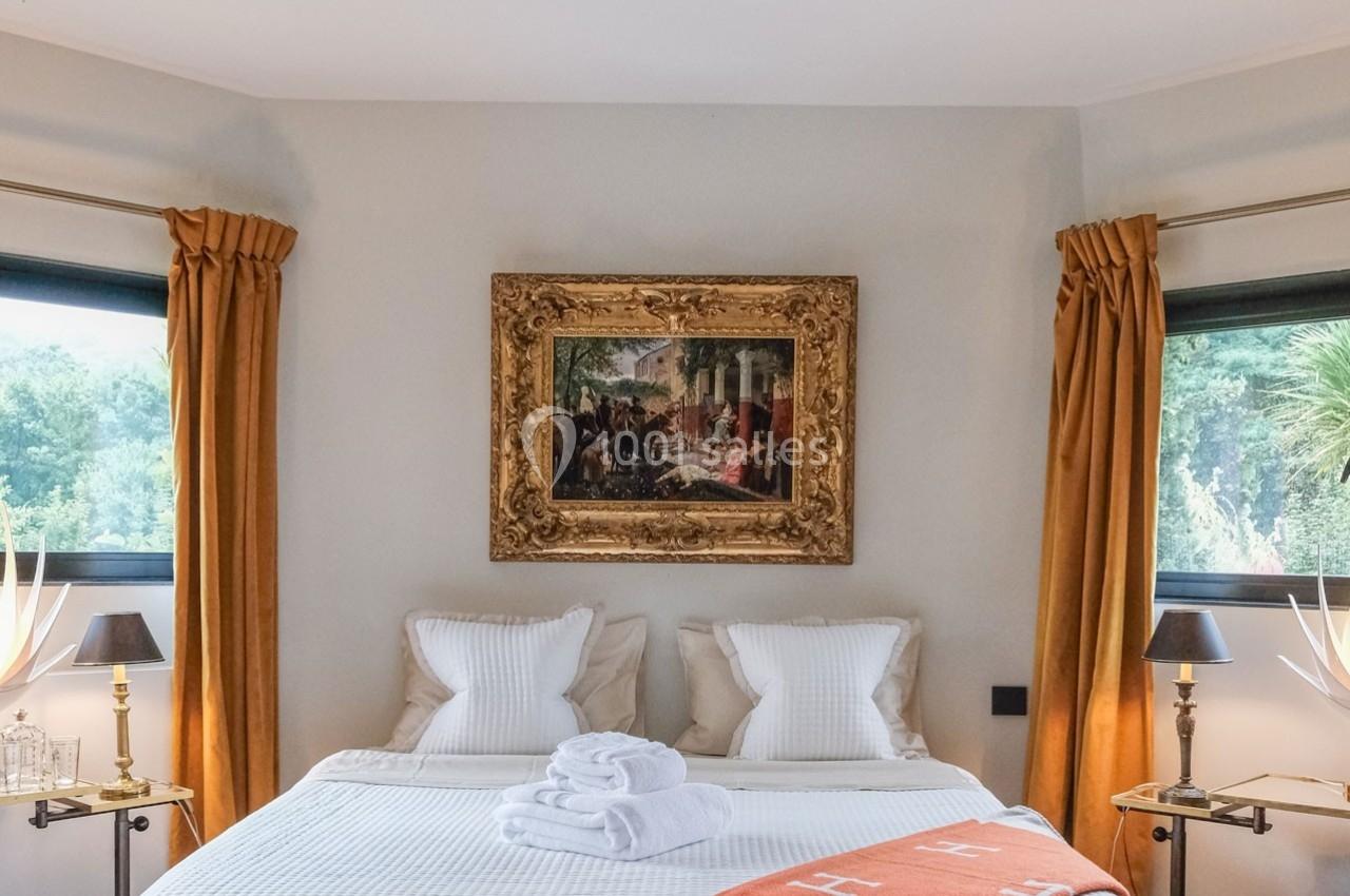 Lit double avec draps blancs, plaid orange, encadré par deux fenêtres et un tableau encadré au mur.