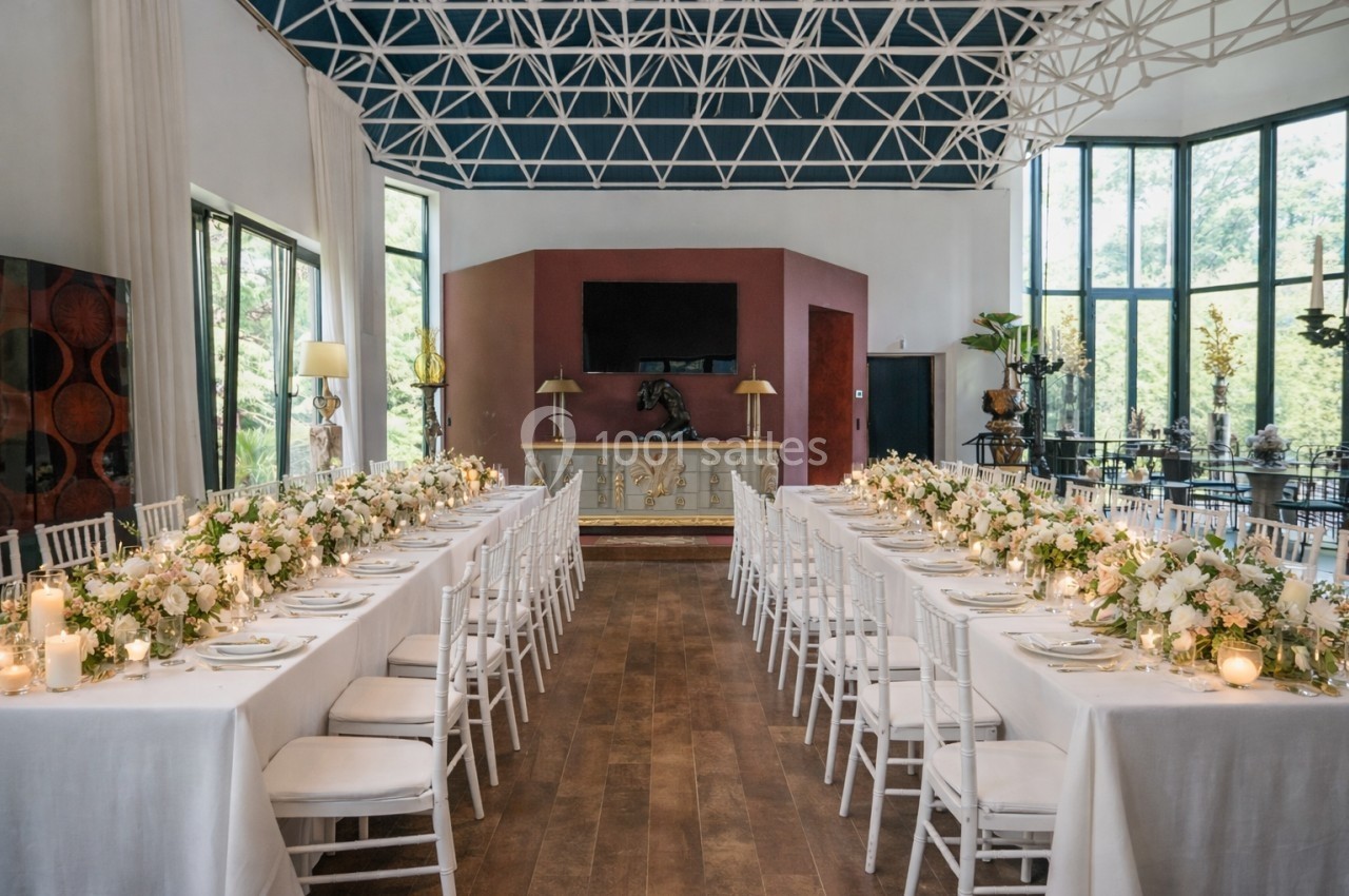 Salle de réception lumineuse avec deux longues tables décorées de fleurs blanches et bougies, entourées de chaises blanches.