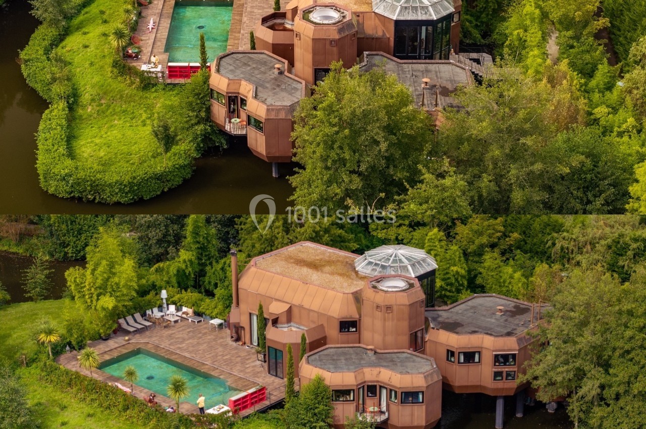Vue aérienne d'une maison moderne entourée d'eau, avec piscine, terrasse et jardin arboré.