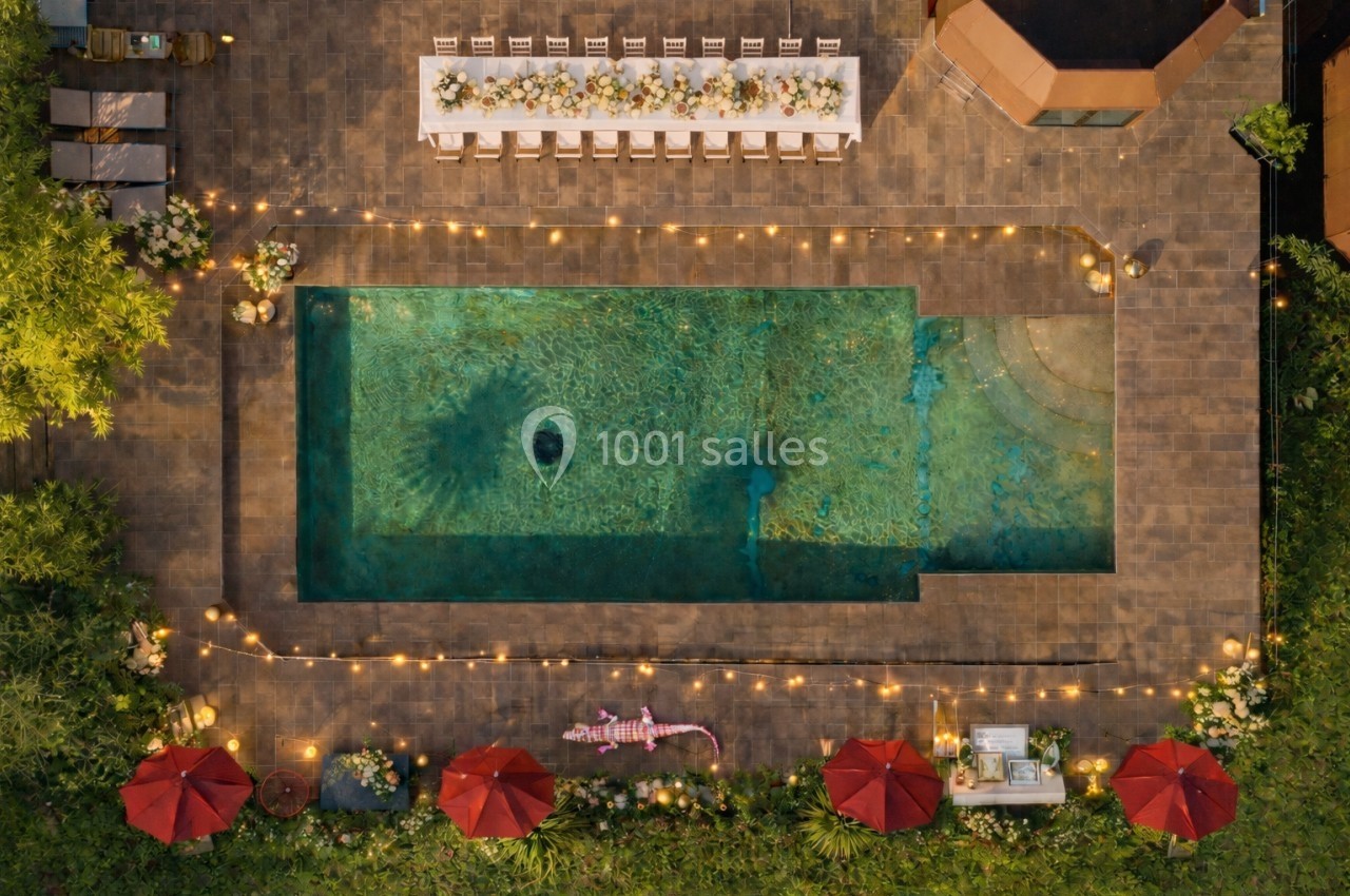 Vue aérienne d'une piscine entourée de guirlandes lumineuses, avec une table dressée et des parasols rouges.