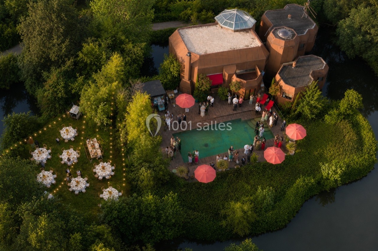 Vue aérienne d'un événement en plein air avec piscine, tables dressées, invités et parasols rouges entourés de verdure.