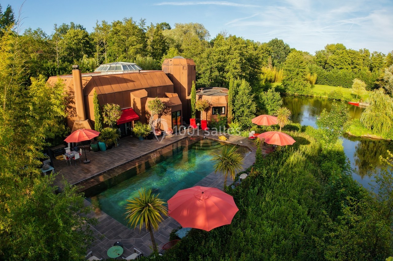 Vue aérienne d'une maison moderne entourée de verdure, avec piscine, terrasses et parasols rouges près d'un étang.