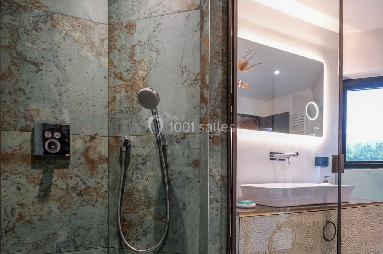 Douche moderne avec paroi vitrée, robinetterie chromée et lavabo en arrière-plan dans une salle de bain lumineuse.