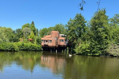 Maison sur pilotis en bois entourée de verdure, située au bord d'un étang calme sous un ciel bleu.