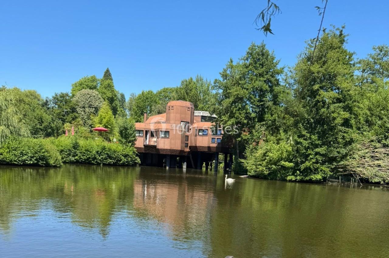Maison sur pilotis en bois entourée de verdure, située au bord d'un étang calme sous un ciel bleu.