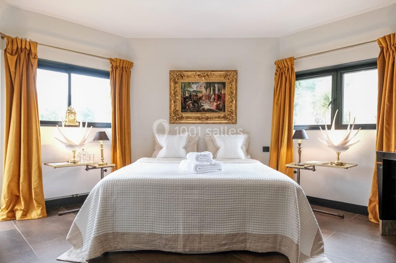 Chambre lumineuse avec lit double, draps blancs, rideaux jaunes, tableaux encadrés et deux fenêtres latérales.