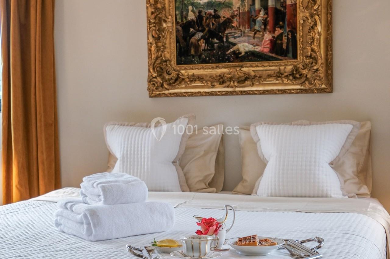 Lit double avec draps blancs, serviettes pliées et plateau de petit-déjeuner, tableau encadré en arrière-plan.