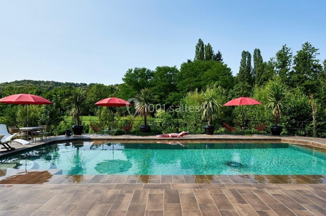 Piscine extérieure avec terrasse en bois, parasols rouges et végétation luxuriante en arrière-plan.