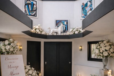 Chambre lumineuse avec lit double, draps blancs, couverture bleue, rideaux turquoise et accès à un balcon.