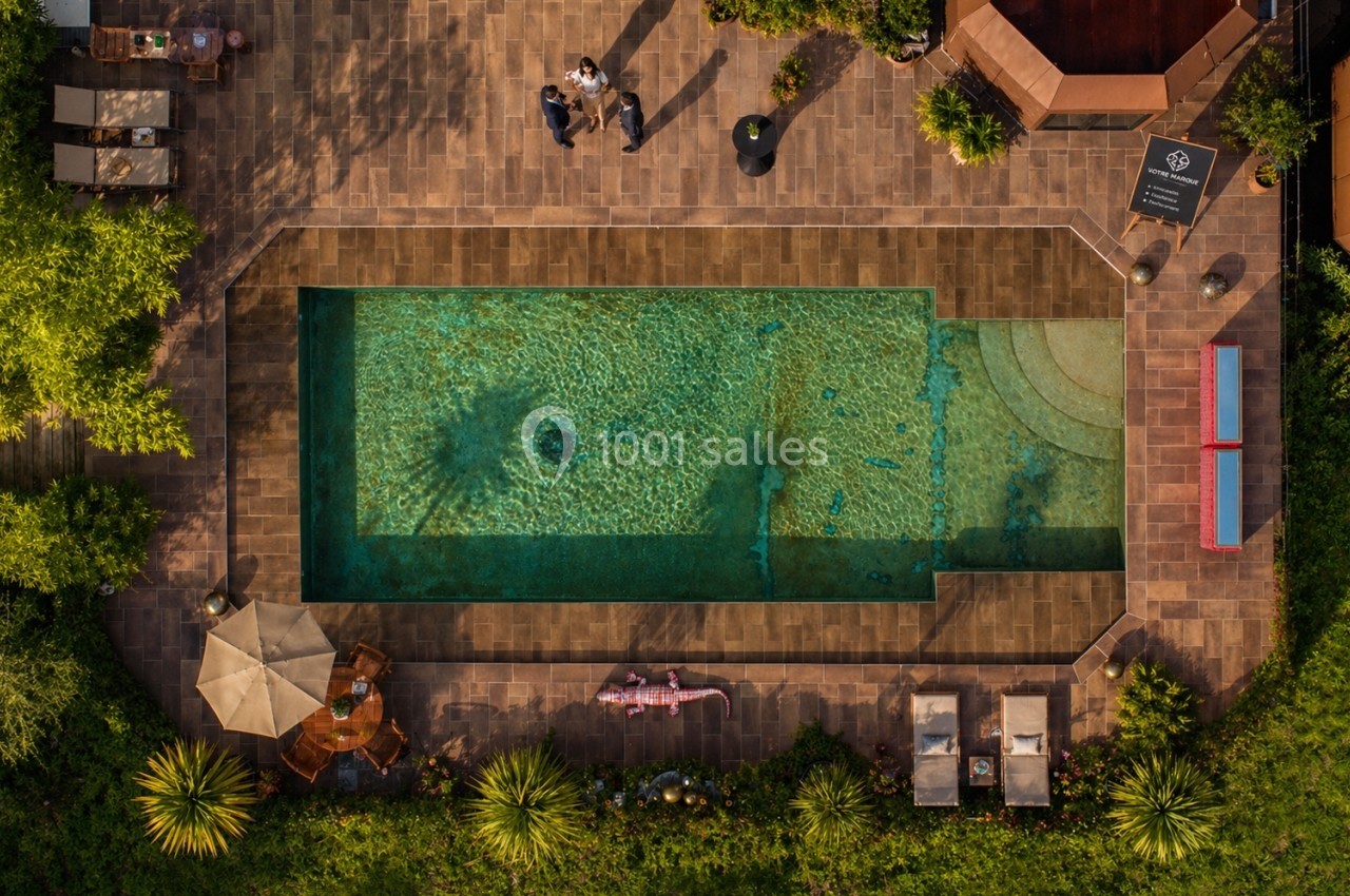 Vue aérienne d'une piscine entourée de terrasses en pierre, de transats, de parasols et de végétation.