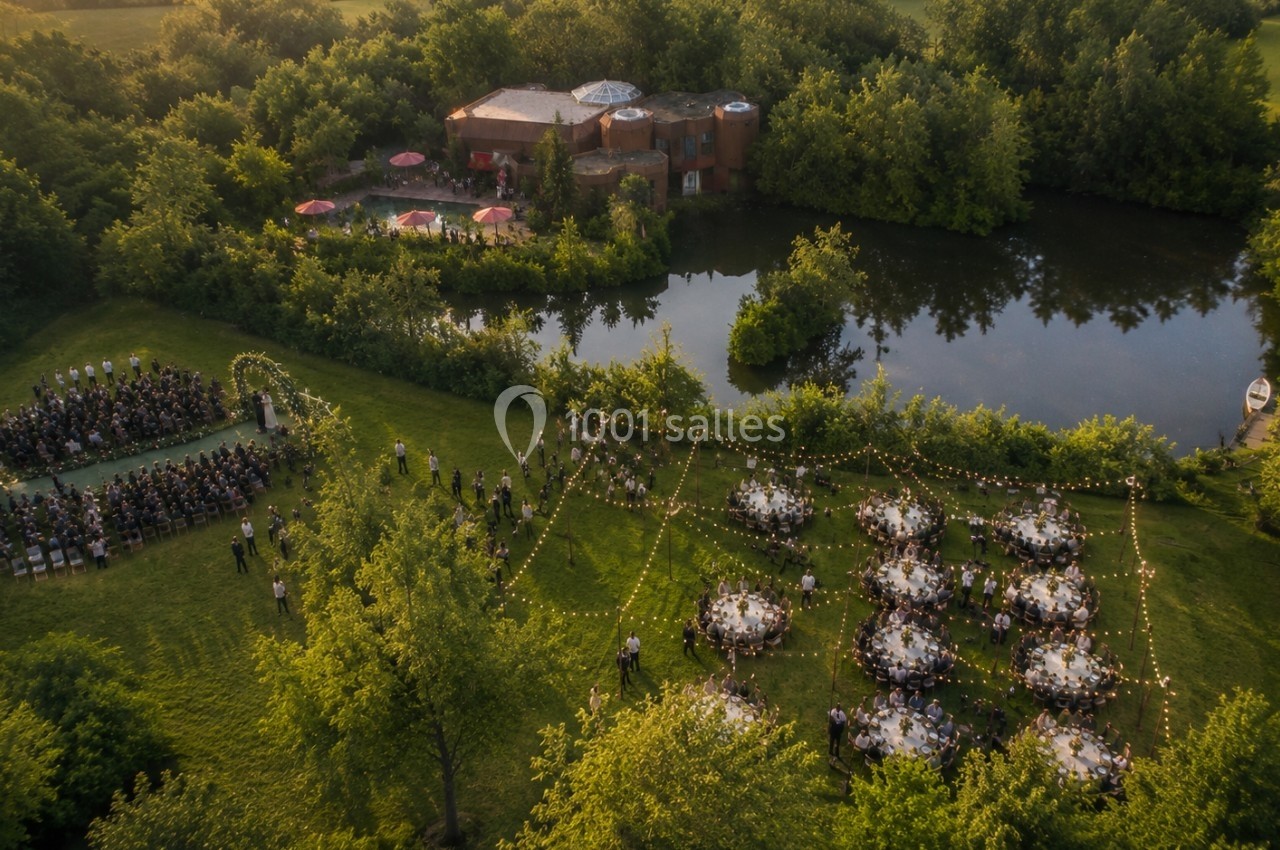 Vue aérienne d'un mariage en plein air avec cérémonie, tables décorées et bâtiment près d'un étang entouré de verdure.